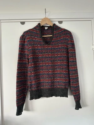 Flerfärgad stickad tröja, 15% mohair- storlek S - Härlig stickad tröja i flera färger med hög V-ringning och långa ärmar, i storlek S (passar även storlek M om man vill ha det mer figurnära). Tröjan har ett randigt mönster i rött, blått, orange, grått och silvertråd/ guldtråd samt ribbstickade muddar och nederkant. 15% mohair som ger materialet en lätt fluffig känsla. Perfekt för dig som gillar en avslappnad och färgglad stil. Mycket gott skick!