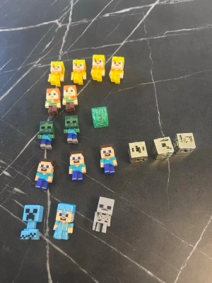 Minecraft minifigurer och block - Samling med små Minecraft-figurer i plast, inklusive karaktärer som Steve, Alex, zombies, creeper, skelett och flera block i guld och grönt. Figurerna är färgglada och fyrkantiga, inspirerade av spelets pixelgrafik. Perfekt för dig som gillar Minecraft och vill ha coola samlarprylar.