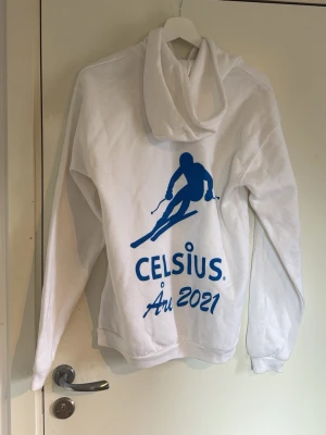 Vit hoodie Celsius Åre 2021 - Vit hoodie med blått tryck av en skidåkare och texten 'Celsius Åre 2021' både fram och bak. Hoodien har huva med dragsko och en stor magficka. Tillverkad i mjukt material, perfekt för chill dagar eller häng med vänner. Storlek M