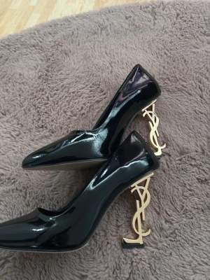 Svarta lackpumps från Saint Laurent - Säljer ett par svarta pumps i glansigt lack från Saint Laurent. Skorna har spetsig tå och unika klackar i guld med YSL-loggan som design. Har knappt använt den. Storleken 39. Har tyvärr ingen  kvitto på det. Men boxen medföljer. Så säljer billigare. Köpte den utomlands 2800kr. Men säljer den för idag 650kr ✨✨🥰🥰 