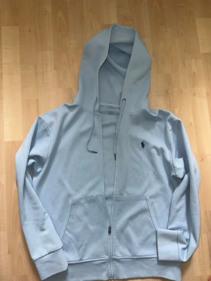 Ljusblå hoodie från Ralph Lauren - Ljusblå hoodie med dragkedja från Ralph Lauren. Klassisk design med huva och dragsnören, två stora fickor framtill och den ikoniska Polo-loggan broderad på bröstet. Tillverkad i mjukt bomullsmaterial som är skönt att ha på sig.