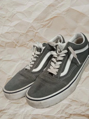 Gråa Vans Old Skool sneakers - Klassiska Vans Old Skool sneakers i grått med vit sula och vit sidostripes. Skorna har lågt skaft, vita snören och detaljer i mocka och canvas. Perfekta för en avslappnad streetwear-look.