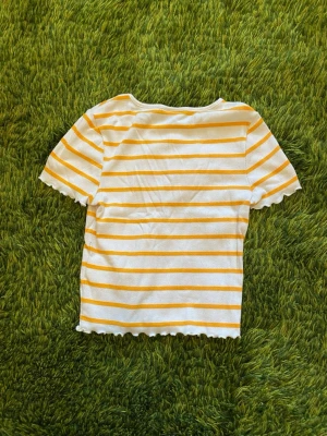 Randig ribbad topp från Gina Tricot - Kortärmad ribbad topp från Gina Tricot i vitt med gula ränder. Har vågiga kanter vid ärmslut och nederkant som ger en söt feminin touch. Passar någon med relativt liten överkropp men tyget är också väldigt stretchigt.