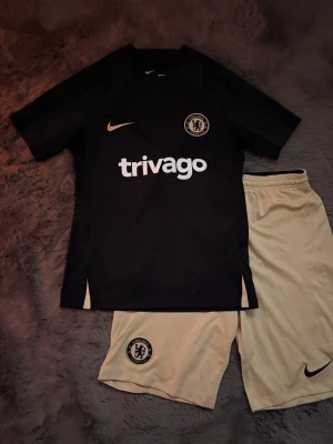 Chelsea träningsset från Nike - Säljer ett Chelsea träningsset från Nike med svart t-shirt och ljusbeige shorts. Tröjan har klubbmärke, Nike-logga och Trivago-tryck på bröstet. Shortsen har elastisk midja och klubbmärke på benet. Perfekt för fotbollsträning eller gym.