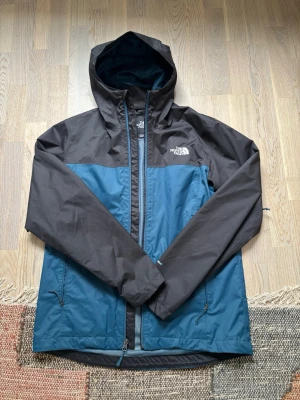 The North Face herrskaljacka  - Snygg och funktionell skaljacka från The North Face i blått och svart. Utrustad med huva, vattentåligt material och två fickor med dragkedja. Perfekt som regn- och vindjacka för vardag, vandring eller friluftsliv. Jackan är i fint skick och alla dragkedjor fungerar som de ska.