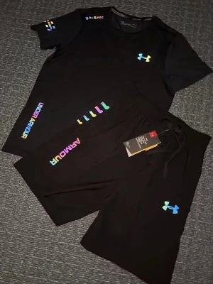 Svart träningsset från Under Armour - Säljer ett svart träningsset från Under Armour med t-shirt och shorts. Setet har färgglada loggor och detaljer i regnbågsfärger. Materialet är mjukt och stretchigt, perfekt för träning eller gym. T-shirten har rund hals och shortsen har dragsko i midjan. Storlek S, helt ny 