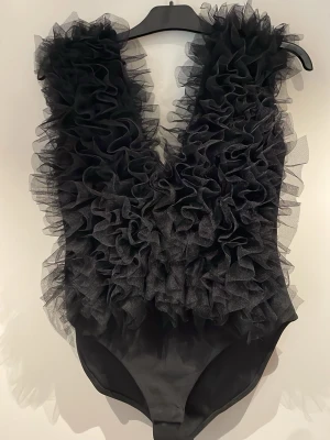 Svart body med volanger i tyll - Supersnygg svart body med djup urringning och massor av volanger i tyll framtill och baktill. Kroppsnära passform och ärmlös design ger en riktigt statement-look. Perfekt för dig som vill sticka ut! Materialet är mjukt och stretchigt, troligtvis polyester.