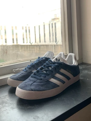 Adidas Gazelle blå sneakers - Snygga Adidas Gazelle sneakers i blå mocka med vita detaljer och klassiska tre ränder på sidorna. Skorna har vit sula och vit hälkappa, samt blå skosnören. Perfekta för en stilren och sportig look.
