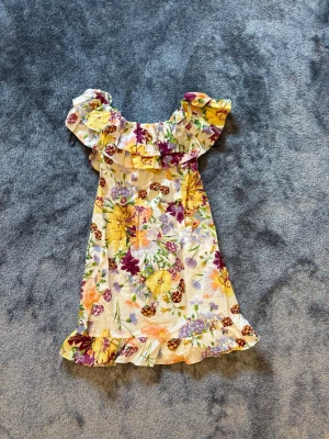 Blommig kortklänning med volang - Söt kortklänning med färgglatt blommönster i gult, lila och orange på vit botten. Klänningen har volangdetaljer vid axlar och fåll samt en luftig passform. Perfekt för varma dagar och somriga tillfällen.