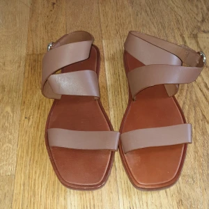 Bruna sandaler med breda remmar - Snygga bruna sandaler med breda remmar i äkta läder och fyrkantig tå. Remmarna korsas över foten och har spänne vid ankeln. Platt sula och stilren design.