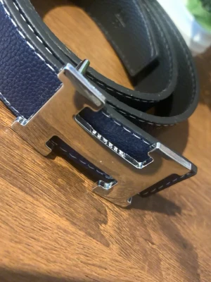 Bälte - Snyggt bälte i mörkblått och svart skinn med ett stort, blankt H-format spänne i silverfärg. Bältet har vit kontrastsöm längs kanterna och klassisk, stilren design som lyfter vilken outfit som helst.