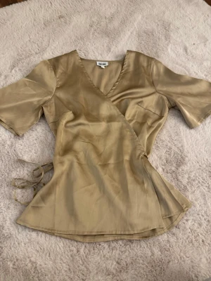 Beige omlottblus - Snygg beige omlottblus från Bubbleroom x Alexandra Nilsson. Blusen har korta ärmar, v-ringning och knytband i sidan. Tillverkad i ett glansigt, satinliknande material som ger en lyxig känsla. Perfekt att styla med jeans eller kjol.
