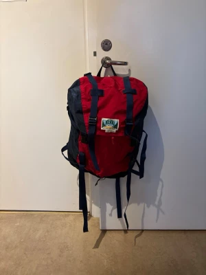 Röd och marinblå ryggsäck Kero - Ryggsäck i rött och marinblått från Kero med justerbara axelband och spännen framtill. Klassisk outdoor-look med stor huvudficka och extra remmar för att fästa utrustning. Tillverkad i slitstark nylon, perfekt för äventyr och vardag.