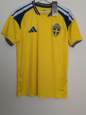 Svensk landslagströja Adidas gul - Säljer en gul svensk landslagströja från Adidas med blå detaljer och Sveriges landslagsemblem på bröstet. Tröjan har korta ärmar, blåa ränder på axlarna och är tillverkad i ett lätt och ventilerande funktionsmaterial. Perfekt för fotbollsträning eller match.