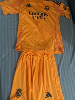 Real Madrid orange matchställ Adidas - Säljer ett orange matchställ från Real Madrid med Adidas-logga och klubbmärke på både tröja och shorts. Tröjan har svarta detaljer, trycket 'Emirates Fly Better' och korta ärmar. Materialet är lätt och andas, perfekt för fotbollsträning eller match.