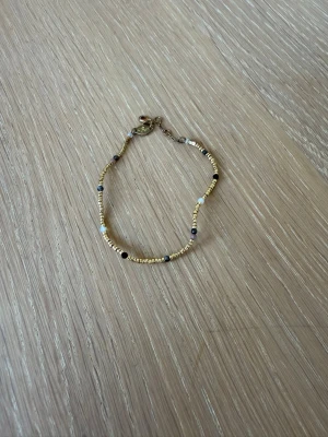 Guldigt pärlarmband med svarta detaljer - Snyggt armband med små guldfärgade pärlor och inslag av svarta och vita pärlor. Armbandet har en enkel och stilren design med metallspänne. Perfekt accessoar för att lyfta din outfit.