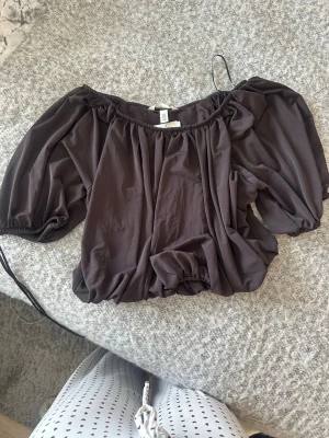 Brun offshoulder blus med puffärm - Säljer en mörkbrun offshoulder blus från H&M med puffiga korta ärmar och dragsko i nederkant. Blusen har en lös passform och är gjord i ett mjukt, tunt polyestertyg som ger ett snyggt fall. Perfekt för dig som gillar trendiga och bekväma toppar.