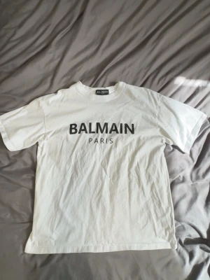 Vit Balmain Paris t-shirt - Snygg vit t-shirt från Balmain Paris med klassisk logga tryckt i svart på bröstet. T-shirten har rund hals och korta ärmar, perfekt för en clean och stilren look. Tillverkad i mjuk bomull för maximal komfort. PASSAR ÄVEN M.