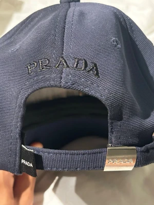 Mörkblå keps från Prada - Snygg mörkblå keps från Prada med broderad logga baktill och metallspänne med ingraverad märkning. Framtill sitter den klassiska trekantiga Prada-plattan i svart och silver. Kepsen är tillverkad i ett slitstarkt tyg med justerbar rem för perfekt passform.