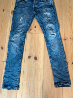 Blå slitna jeans från Jack and Jones  - Säljer ett par blå slim fit/skinny jeans med slitningar och trasiga detaljer på låren och knäna. Jeansen har klassisk femficksmodell och för skön passform. Perfekta för en avslappnad och trendig look.