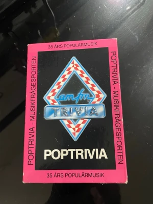 Poptrivia musikfrågesport spel - Poptrivia är ett frågesportspel om 35 års populärmusik. Spelet innehåller kort med frågor om olika musikgenrer som rock, pop, soul och modern rock. Lådan är rosa och svart med färgglada spelkort i blått, rött och vitt.