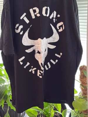 Svart t-shirt med tjurtryck 3XL - Cool svart t-shirt i storlek 3XL med stort vitt tryck av en tjurskalle och texten 'STRONG LIKE BULL' på framsidan. T-shirten har rund hals och korta ärmar. Perfekt för dig som gillar statement-plagg och streetwear vibes.
