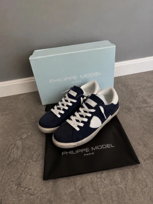 Philippe Model Low PRSX Mörkblå Suede - Snygga sneakers från Philippe Model i mörkblå mocka med vita detaljer och vita skosnören. Skorna har en klassisk låg siluett och vit sula. Perfekta för dig som gillar stilrena och trendiga sneakers med premiumkänsla. Nypris : 2800kr. Ny skick. Skorna har suede material. Box och Dustbag tillkommer. 