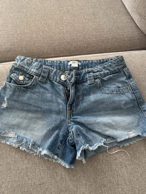 Blå jeansshorts från Ginatricot - Säljer ett par blå jeansshorts från Ginatricot med fransig kant och slitna detaljer. Klassisk femficksmodell med knapp och dragkedja framtill. Perfekta för varma dagar och enkel att matcha med olika toppar.