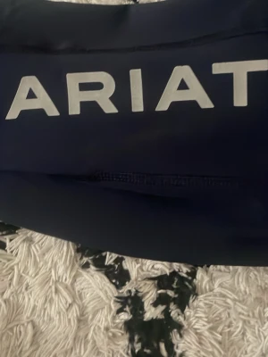 Mörkblå ridbyxor från Ariat - Snygg mörkblåa ridbyxor från Ariat med stort vitt logotryck på låret. Tillverkad i ett stretchigt syntetmaterial som andas, perfekt för aktiva dagar. Modellen är tight och har en sportig look med diskreta sömmar.