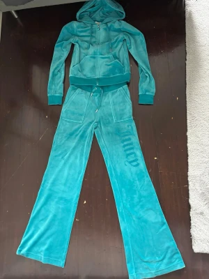 Turkos Juicy Couture velourset - Juicy Couture loungewear-set i mjuk turkos velour. Setet består av en zip-hoodie med fickor och huva samt matchande byxor med dragsko och fickor. Både jackan och byxorna har Juicy-logga i strass. Endast använd ett fåtal gånger men armen har ett hål på sig men när man har på sig den tänker man knappt på det.