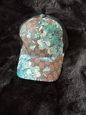Gucci keps med blått blommönster - Snygg keps från Gucci med klassiskt GG-mönster i brunt och ett färgstarkt blommönster i blått, turkos och vitt. Kepsen har svart mesh baktill och justerbar rem. Perfekt för dig som vill sticka ut med en exklusiv accessoar.