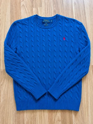 Blå kabelstickad tröja från Polo Ralph Lauren - Snygg blå kabelstickad tröja från Polo Ralph Lauren med rund halsringning och röd broderad logga på bröstet. Tröjan har långa ärmar och ribbade muddar vid ärmslut och nederkant. Perfekt för en stilren och klassisk look.