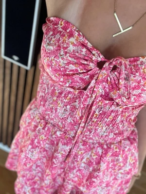 Rosa blommig axelbandslös klänning - Söt kortklänning i rosa med blommigt mönster och smockad midja. Axelbandslös med snörning framtill och volangdetaljer. Endast använd fåtal gånger💓Köpt utomlands