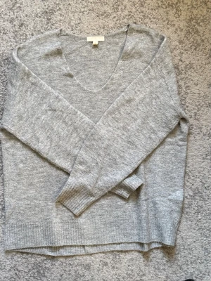 Ljusgrå stickad tröja från H&M - Mysig ljusgrå stickad tröja från H&M med v-ringning och långa ärmar. Tröjan har en avslappnad passform och ribbade muddar vid ärmslut och nederkant. Perfekt att slänga på sig när det är kyligt ute.