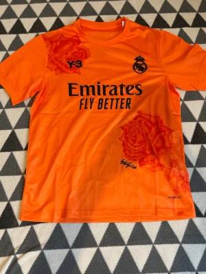 Orange Real Madrid tröja Adidas S - Snygg orange Real Madrid matchtröja från Adidas med svarta tryck och röda rosdetaljer. Tröjan har korta ärmar, rund hals och är tillverkad i lätt funktionsmaterial (AEROREADY). Perfekt för fotboll eller träning.