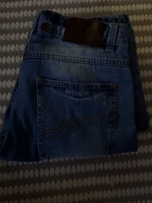 Blå raka jeans med läderpatch - Snygga blå jeans med klassisk femficksdesign och raka ben. Jeansen har en mörk läderpatch bak i midjan och subtila slitningar för en avslappnad look. Tillverkade i denim, perfekta för en casual stil.