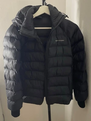 Svart dunjacka Peak Performance - Snygg svart dunjacka från Peak Performance med huva och dragkedja framtill. Jackan har puffig design, liten logga på bröstet och muddar vid ärmsluten. Perfekt för kalla vinterdagar och riktigt varm tack vare dunfyllningen.
