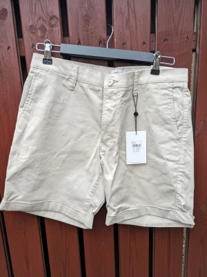 Oanvända These Glory Days, beige shorts, storlek medium - Oanvända (inkl. prislapp) korta shorts i chinosvariant från These Glory Days, modellen T Aimon Shorts (slim) i bomullstwill med en ljusbeige färg. Shortsen har en klassisk design med fickor fram och bak samt hällor för bälte. Uppviket är fastsytt av tillverkaren. De är försedda med en knapp och dragkedja framtill. Säljs eftersom de inte kommit till användning då de helt enkelt är för tajta för min kropp. Är som referens 170 cm lång och väger 72-73 kg.   Midja: 83 cm.  Benlängd: C:a 40 cm.