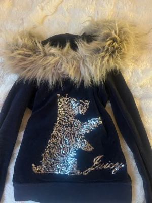 Juicy Couture hoodie med paljetter & fuskpäls - Mörkblå hoodie från Juicy Couture med dragkedja, stor huva med fluffig fuskpäls och glittrig paljettdekor i form av en hund på ryggen. Mjuk velourkänsla och långärmad modell. Perfekt statement-plagg för dig som gillar bling och cozy vibes. Storlek L men passar S bättre. 
