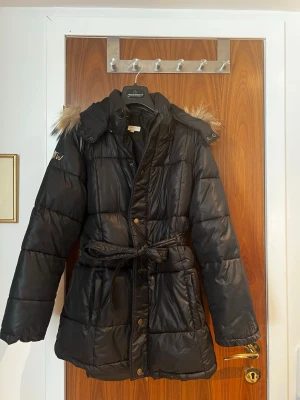 Svart pufferjacka med fuskpäls - Snygg svart pufferjacka från Tina Wodstrup med avtagbar huva och fuskpälsdetalj. Jackan har bälte i midjan för en schysst siluett och stängs med både dragkedja och knappar. Perfekt för kalla vinterdagar och riktigt varm.