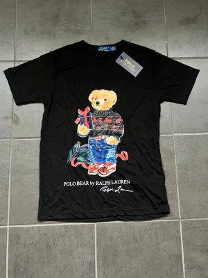 Svart Polo Bear t-shirt från Ralph Lauren - Snygg svart t-shirt från Ralph Lauren med ikoniska Polo Bear-trycket framtill. Björnen har en julig tröja, jeans och håller ett paket med röd rosett. T-shirten har rund hals och korta ärmar. Perfekt statement-plagg för dig som gillar klassiska motiv. Jag kan också skicka den samma dag👍