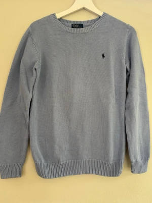 Ljusblå stickad tröja Polo Ralph Lauren - Snygg ljusblå stickad tröja från Polo Ralph Lauren med klassisk rund halsringning och svart broderad logga på bröstet. Tröjan har ribbade muddar vid ärmslut och nederkant. Perfekt för lager på lager och enkel att matcha.