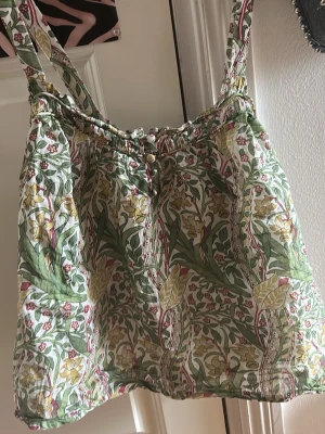 Blommig linnetopp från Morris & Co - Supersöt linnetopp från Morris & Co x Next med grönt, gult och rött blommönster. Toppen har smala axelband, knappar framtill och är gjord i tunn bomull som känns luftig och perfekt för varma dagar. Perfekt att styla med jeans eller kjol!