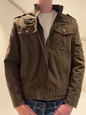Olivgrön utilityjacka - Snygg olivgrön utilityjacka med framfickor, axelklaffar och knappar. Jackan har dragkedja framtill samt detaljer i militärstil. Perfekt för dig som gillar en avslappnad och cool look.