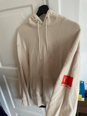 Beige hoodie från Calvin Klein Jeans - Snygg beige hoodie från Calvin Klein Jeans med stor CK-logga och röd text på ryggen samt röd logga på ärmen. Hoodien har huva med dragsko och en klassisk känguruficka framtill. Tillverkad i mjukt bomullsmaterial för skön känsla.