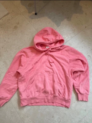 Coral/rosa hoodie med huva - Säljer en klassisk coral/rosa hoodie med huva och dragsko. Hoodien har en stor magficka framtill och ribbade muddar vid ärmslut och nederkant. Perfekt för chill dagar eller när du vill ha en enkel och snygg look. Materialet känns mjukt och bekvämt. Den har små maskara fläckar på ena ärmen men går bort i tvätten.