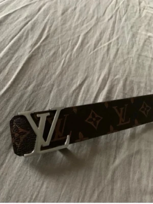  monogram bälte - Snyggt bälte  med klassiskt monogrammönster i brunt och guld. Bältet har en stor logga i silver som spänne och är gjort i skinn. Perfekt accessoar för att lyfta vilken outfit som helst.