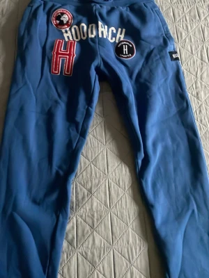 HOODRICH BYXOR BLÅ - Säljer ett par blå mjukisbyxor från Hoodrich med vita och röda detaljer samt broderade patches och loggor. Byxorna har dragsko i midjan och är tillverkade i mjukt material som känns skönt mot huden. Perfekta för en avslappnad och trendig stil.