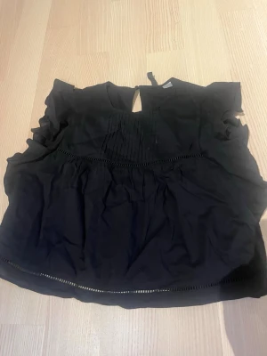 Svart ärmlös blus med volang - Snygg svart blus med volangdetaljer vid ärmhålen och dekorativa veck framtill. Blusen har rund halsringning med liten öppning och knapp bak i nacken. Materialet ser ut att vara lätt och luftigt, perfekt för en trendig look.
