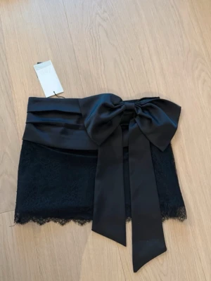 Svart kortkjol med stor rosett - Supersnygg svart kortkjol med bred satinmidja och en stor dekorativ rosett framtill. Kjolen har spetsdetaljer nedtill som ger en elegant touch. Perfekt för dig som vill sticka ut med en unik och trendig look. Lappen sitter inte kvar men aldrig använd😊